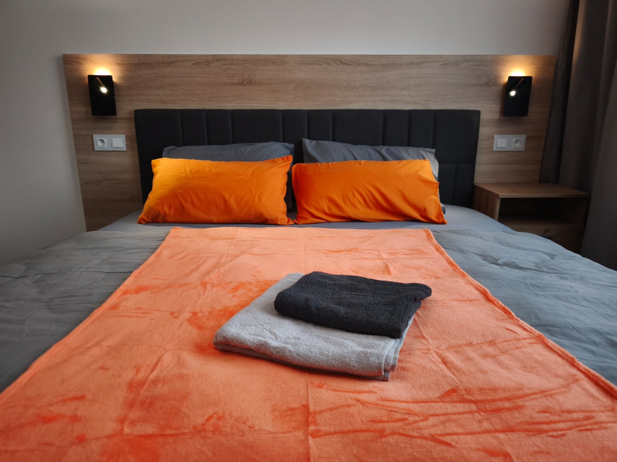 A comfortable sleeping area is presented, featuring a spacious bed with dark grey and bright orange pillows. An orange throw blanket is laid out over the bed, with neatly folded towels in gray and black positioned on top.