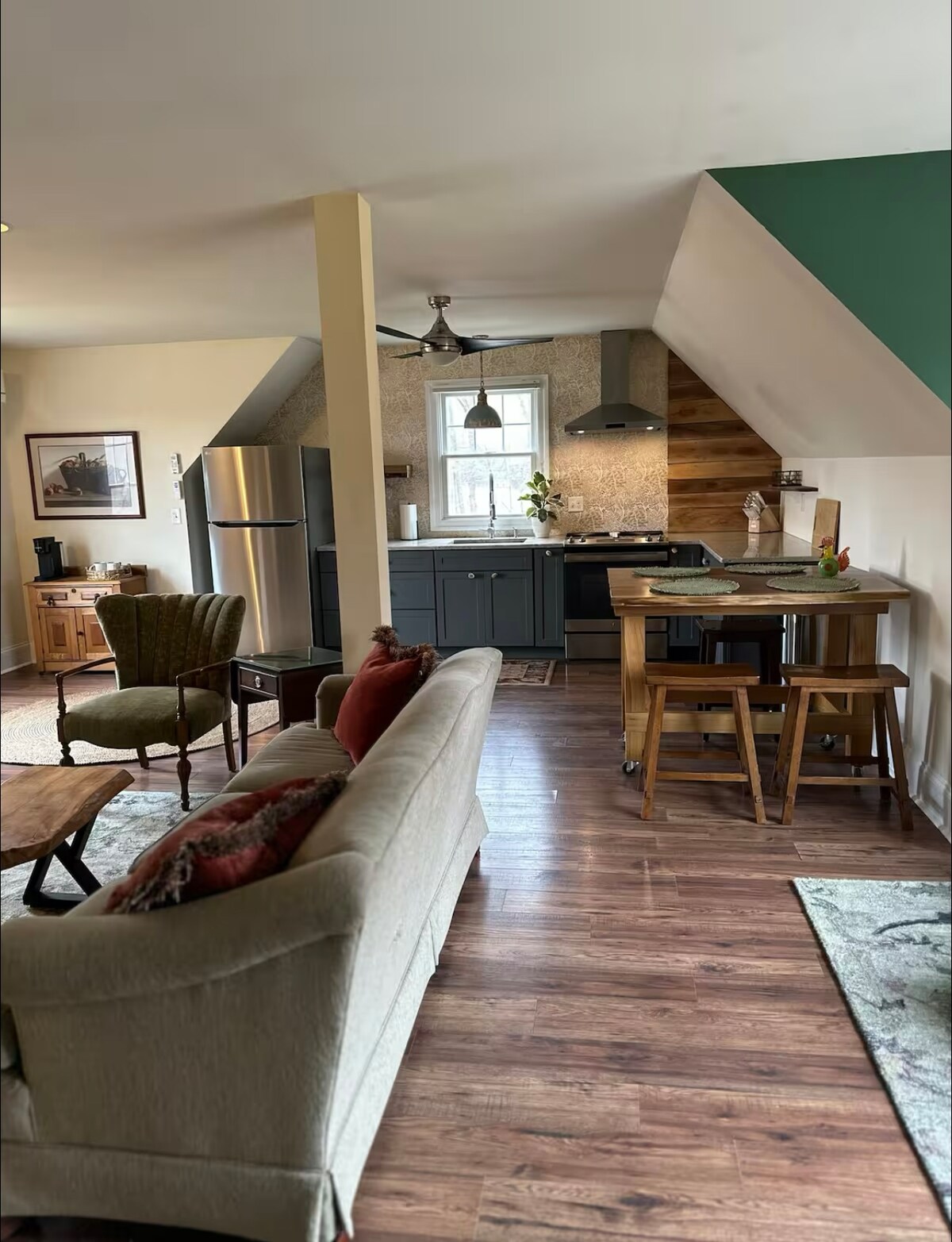 The open living space features a cozy seating area with a light-colored sofa and an accent chair. The kitchen is equipped with modern appliances, including a refrigerator and stove. A dining table with two stools occupies a corner, blending seamlessly with warm wooden accents.