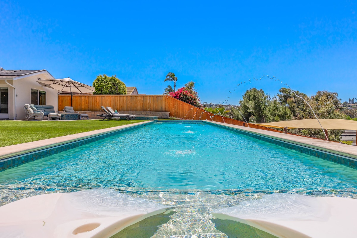 A long, shimmering saltwater lap pool stretches towards a clear blue sky, surrounded by a grassy area and well-maintained landscaping. Lounge chairs line one side, while a covered patio sits in the background, providing space for relaxation and outdoor enjoyment.