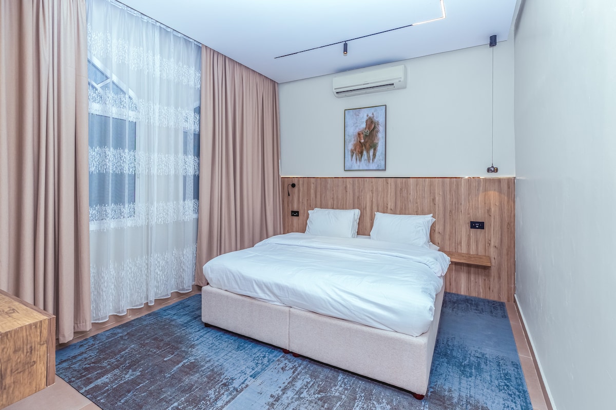 A cozy sleeping area is presented, featuring a spacious bed with crisp white bedding. Natural light filters through a sheer curtain, illuminating the wooden accents on the walls. A piece of artwork complements the warm tone of the room, enhancing the overall modern decor.