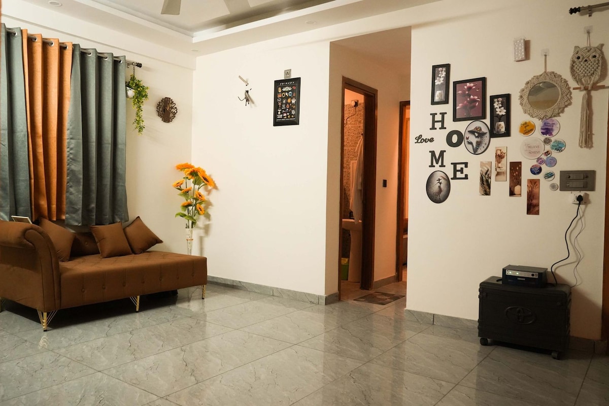 A modern living area features a brown upholstered sofa with decorative cushions. Wall decor includes framed artwork and a vibrant 'HOME' display. A potted plant adds a touch of greenery, while the tiled floor reflects soft lighting from above.