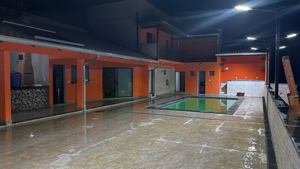 An illuminated outdoor space is showcased at night, featuring a pool surrounded by a spacious tiled area. The vibrant orange walls of the surrounding structure create contrast against the dark sky, with several outdoor lights providing additional visibility.