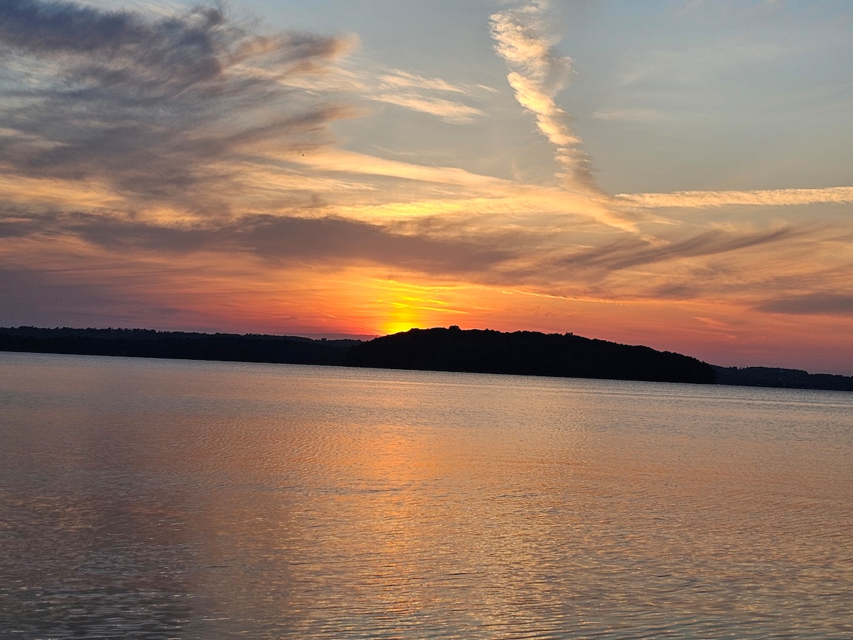A serene sunset over Rice Lake, showcasing vibrant hues of orange, pink, and blue reflecting off the calm water. A distant island is visible on the horizon, framed by gentle clouds that enhance the tranquil atmosphere.