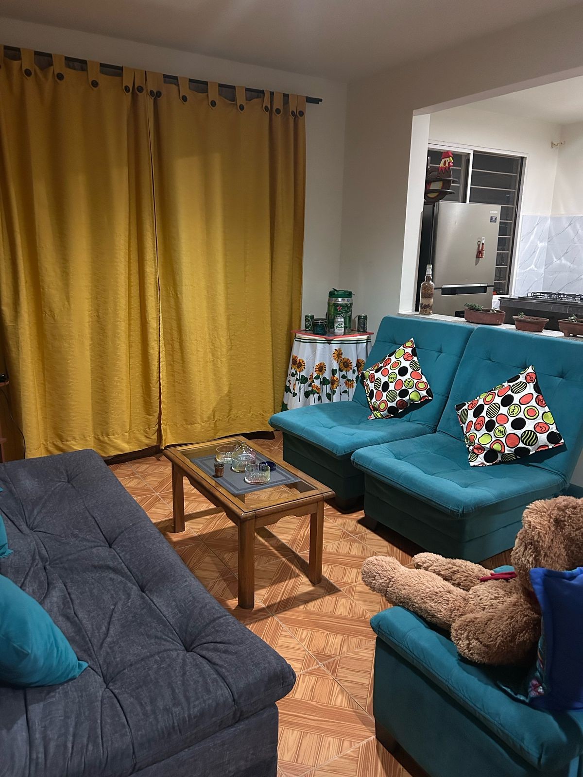 A cozy living area is presented, featuring a gray sofa and two teal chairs with floral-patterned cushions. A wooden coffee table sits in front, complemented by a bear plush toy. Bright yellow curtains frame the room, adding a splash of color, while a glimpse of the kitchen is visible in the background.