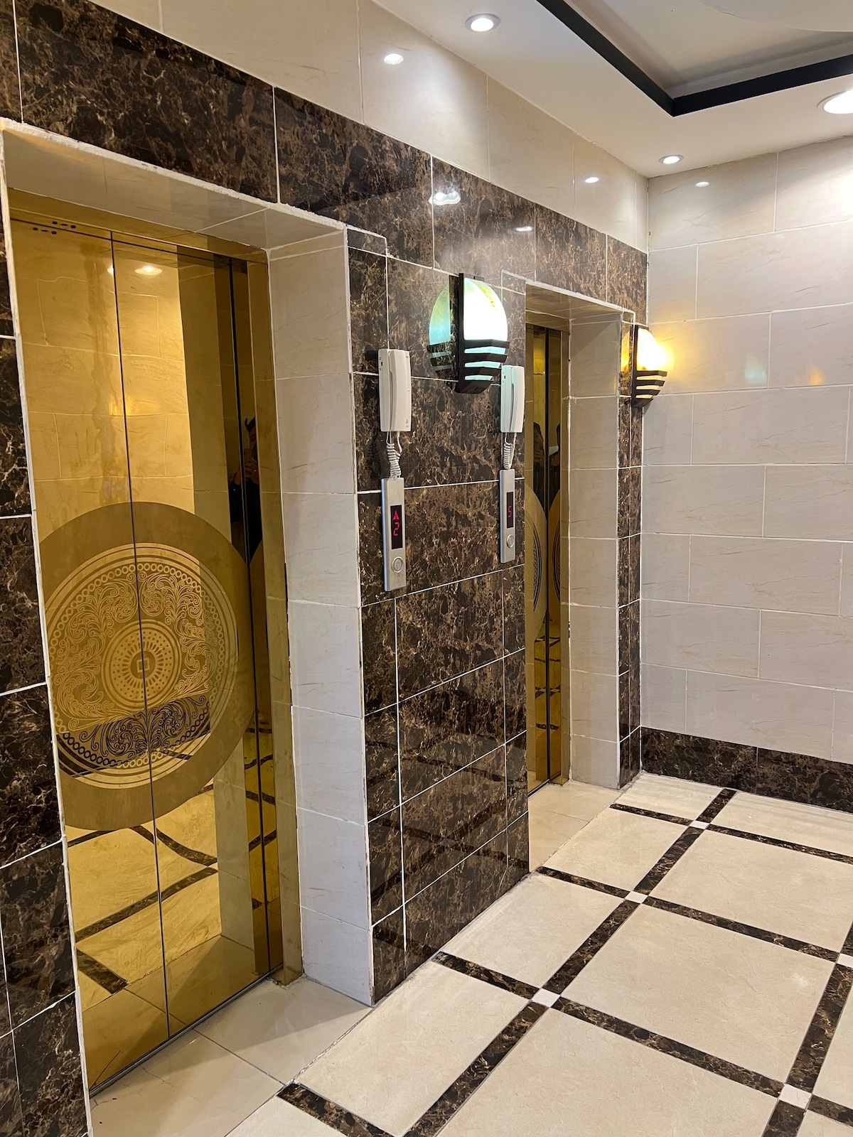 A hallway is displayed featuring two elegant glass elevators set against polished marble walls. Soft lighting enhances the warm hues of the decor. Floor tiles are arranged in a contrasting pattern, creating a sophisticated and inviting entryway.