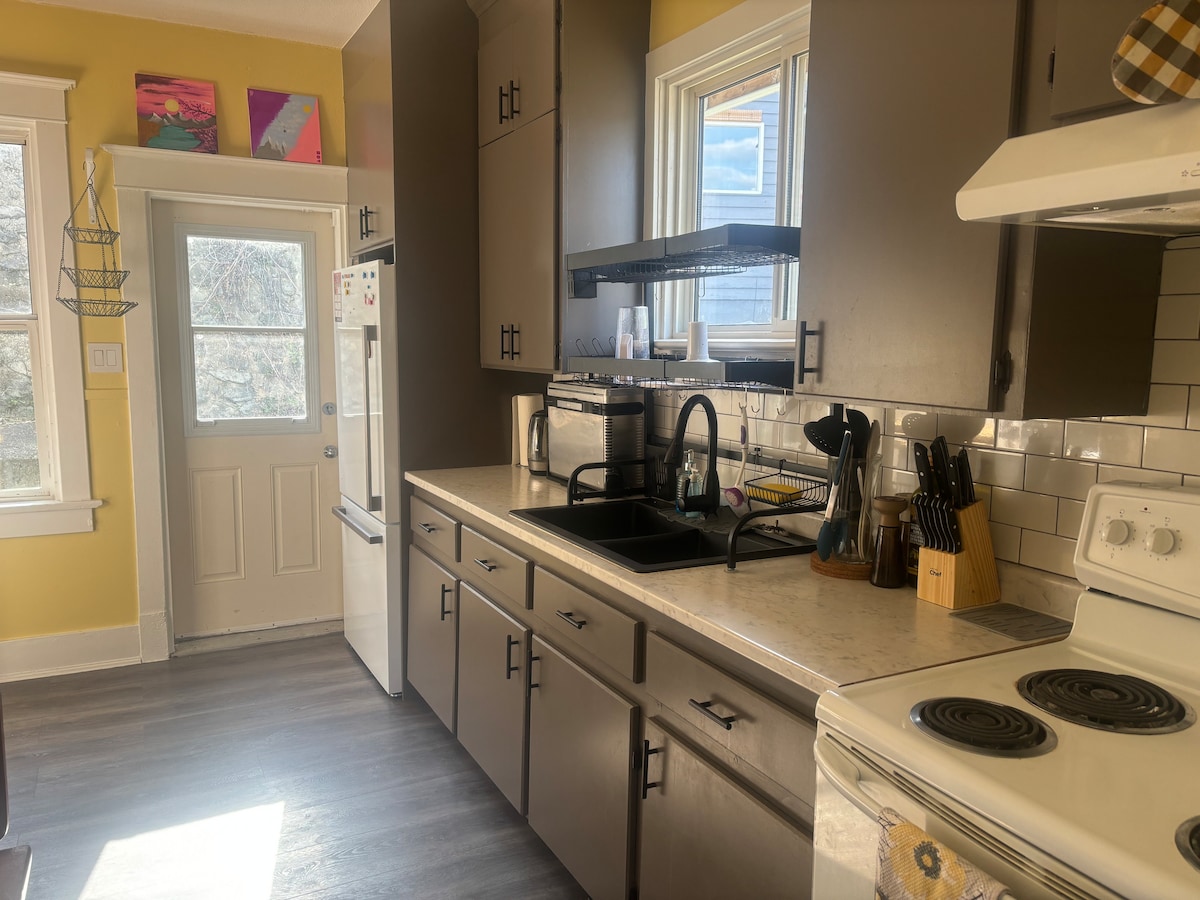 A well-organized kitchen features light-colored cabinetry and a spacious countertop. A sink is situated near a window, allowing natural light to enhance the space. Kitchen utensils and appliances are neatly arranged, creating a functional area for meal preparation.