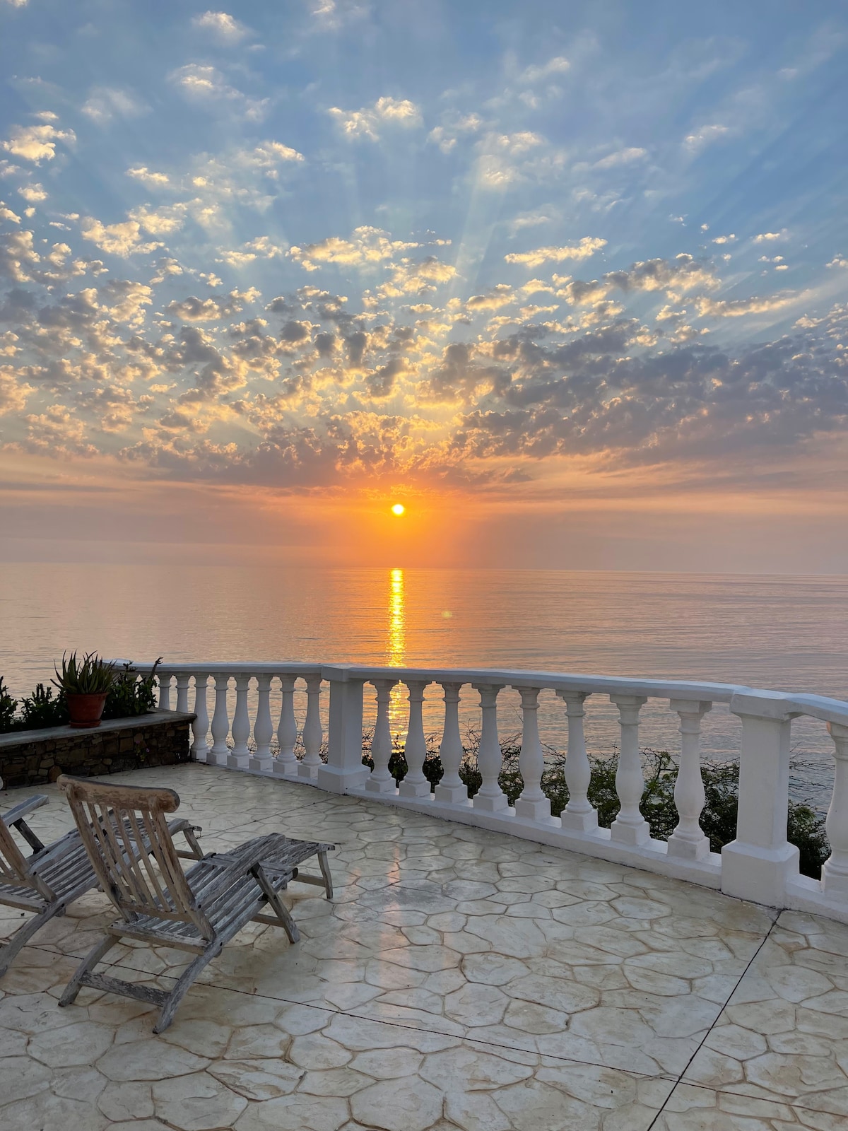 An inviting terrace is showcased, featuring two wooden lounge chairs facing a vibrant sunset over the calm sea. The sky is filled with soft, multicolored clouds, reflecting warm light that bathes the terrace. A stone railing borders the space, providing a sense of openness.