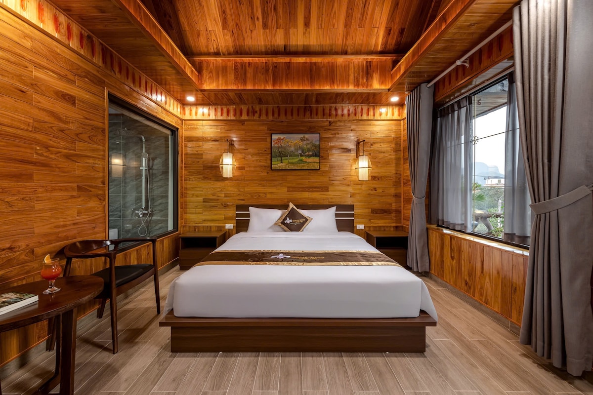 A cozy room with warm wood paneling and a spacious bed is illuminated by soft lighting. A large window offers views of the outside, while a small table and two chairs provide seating. A painting hangs on the wall, adding character to the space.