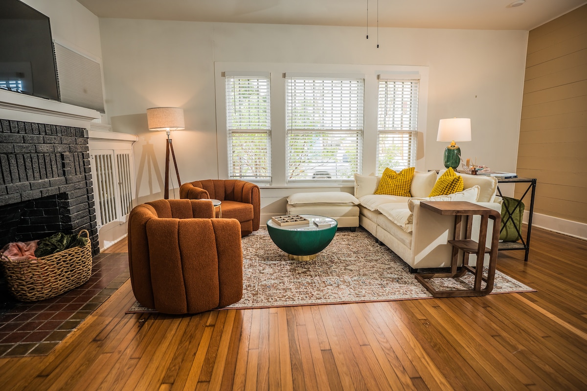 A cozy living area features two curved orange armchairs and a white sectional sofa, arranged around a coffee table and area rug. Natural light filters through the large windows, creating a warm ambiance. A floor lamp and decorative items add to the inviting atmosphere.