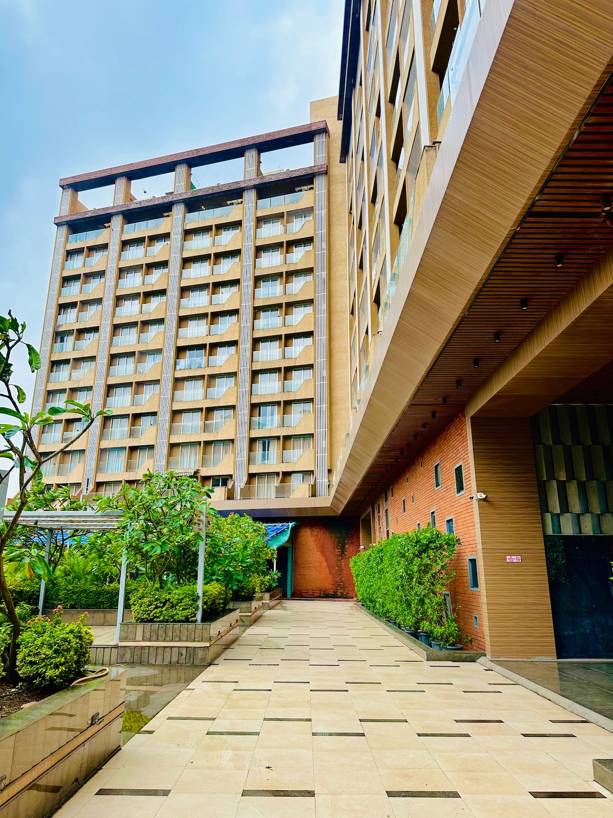A modern hotel building is observed, featuring a combination of glass and textured facades. Lush greenery frames the walkway, which is decorated with square tiles. The structure's upper levels display rows of windows, allowing natural light into the interior spaces.