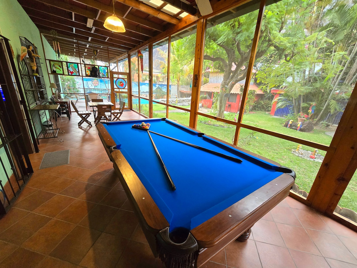 A spacious game room features a blue felt pool table set against large glass windows that allow natural light to fill the area. Lush greenery from the garden is visible outside, creating a relaxed and inviting atmosphere. A cozy seating arrangement complements the space.