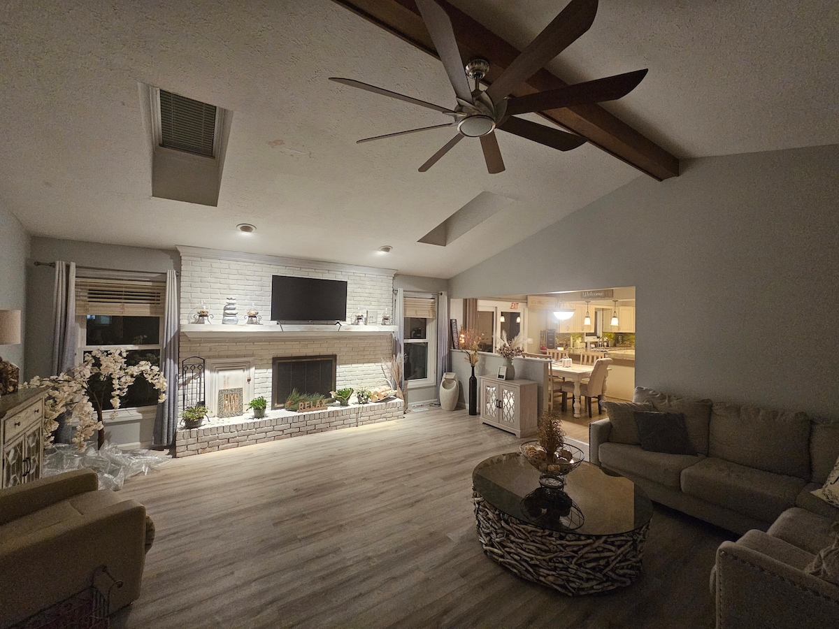 A spacious living room is highlighted by a vaulted ceiling with wooden beams. A large flat-screen television is mounted above the fireplace, surrounded by decorative plants. Soft seating options include a neutral-toned sectional and wicker coffee table, while natural light filters through the windows.