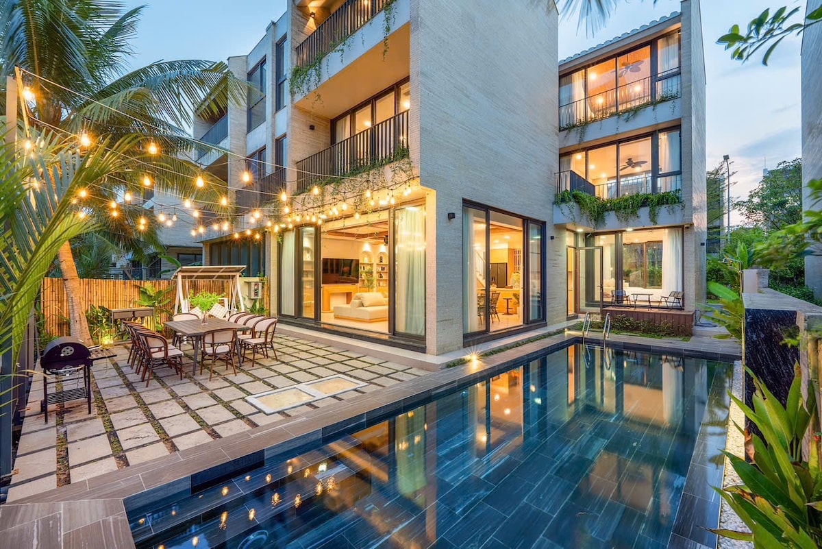 A spacious outdoor area features a private swimming pool reflecting the evening sky. Lounge seating and a dining table are arranged under decorative string lights. The villa's modern architecture is visible, with large glass doors opening to the interior, surrounded by lush greenery.