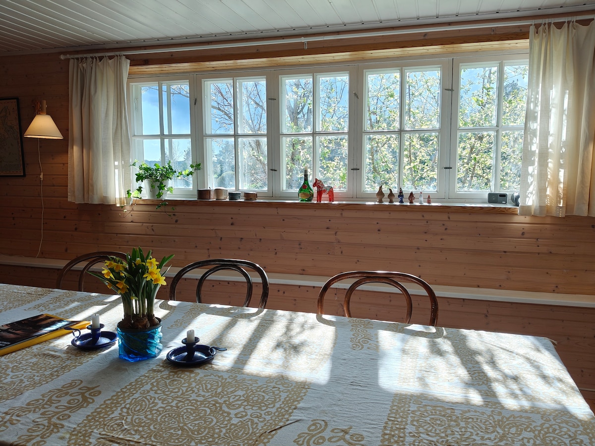 A spacious dining area features a long table adorned with a light-colored tablecloth. Natural light floods in through multiple windows, revealing a view of greenery outside. Decorative items are arranged on the windowsill, complemented by a small potted plant and a bouquet of flowers.
