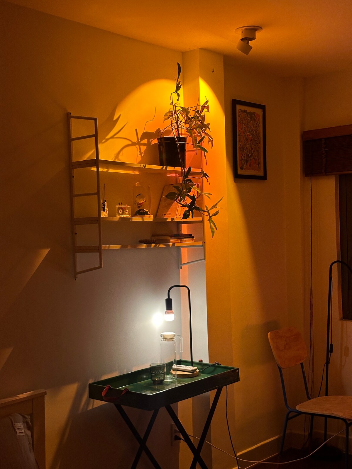 A well-lit corner features a small table with a glass of water and a plant on top. Wall-mounted shelves display decorative items and a framed artwork is hung nearby. Soft orange lighting creates a calming ambiance, highlighting the textures and shapes in the space.