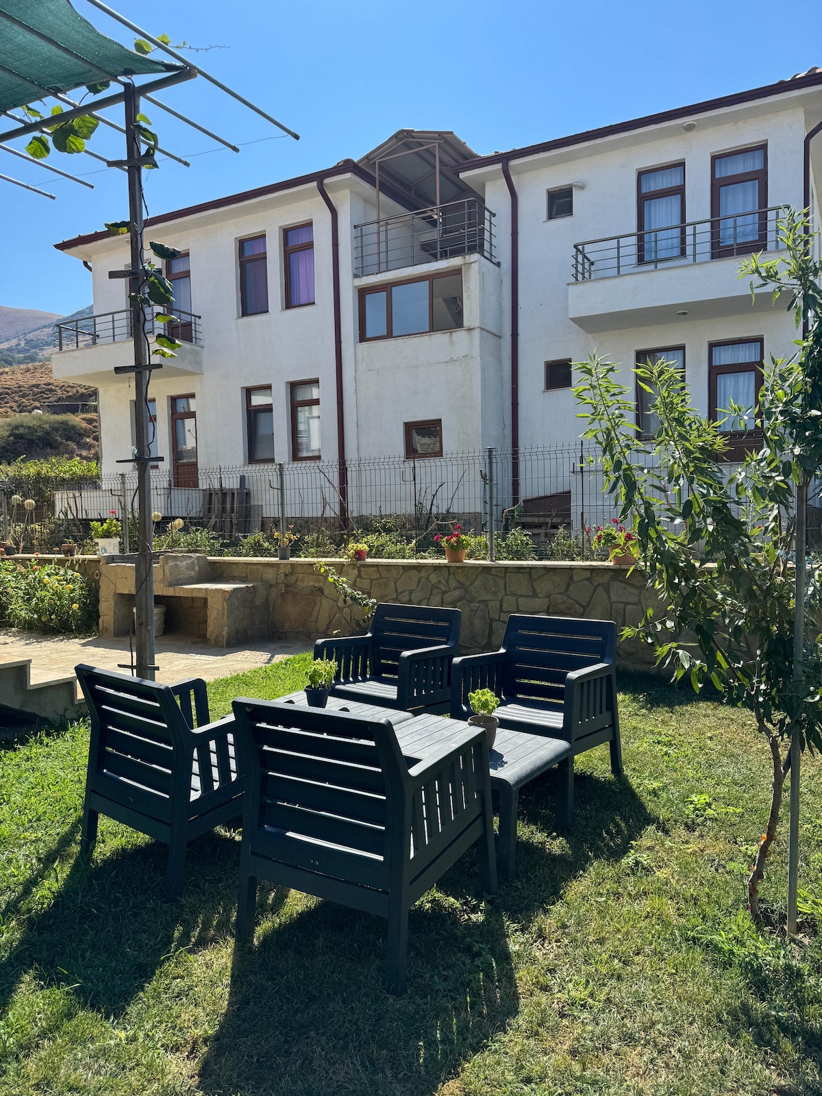 A spacious outdoor area features a set of four black wooden chairs arranged around a small table on a grassy lawn. The backdrop consists of a multi-story building with large windows and a balcony. A gardening trellis with climbing plants is visible in the scene.