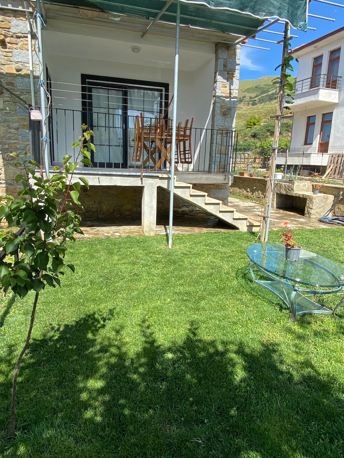 A private garden area is visible, featuring well-maintained grass. A wooden dining table with chairs is positioned on a small patio, accessible by a few steps. A glass table with a potted plant sits nearby, complemented by the stonework of the building.