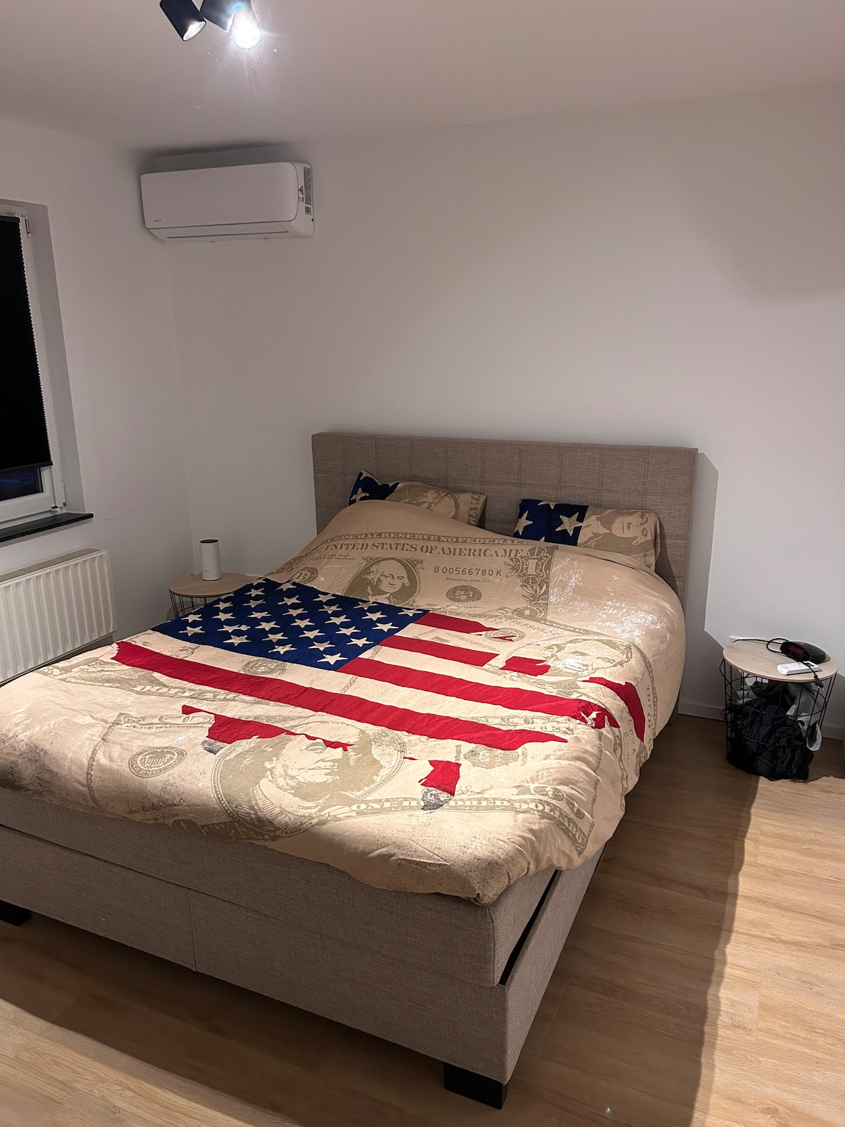 A spacious bed is adorned with a flag-themed comforter, complemented by decorative pillows. The light-toned walls and wooden flooring create a bright and airy ambiance. An air conditioning unit is mounted on the wall, and a small nightstand is positioned beside the bed.