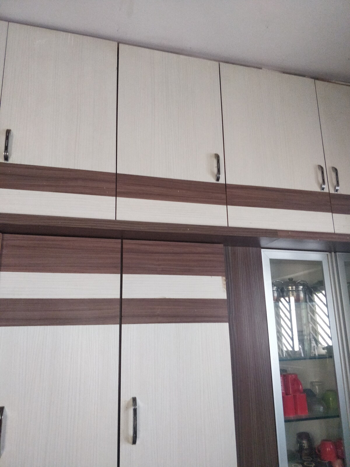 A modern cabinet is shown with a mix of light and dark wood paneling. The upper section features several sleek, silver handles, while the lower section includes glass doors that allow visibility to the items inside, enhancing the storage space.