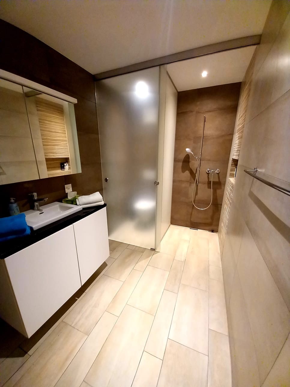 A modern bathroom features a glass-enclosed shower with a sleek wall-mounted shower head. A minimalist vanity with a dark countertop and storage space is seen, accompanied by neatly arranged towels. Warm, neutral tones and large tiles create a clean, open atmosphere.