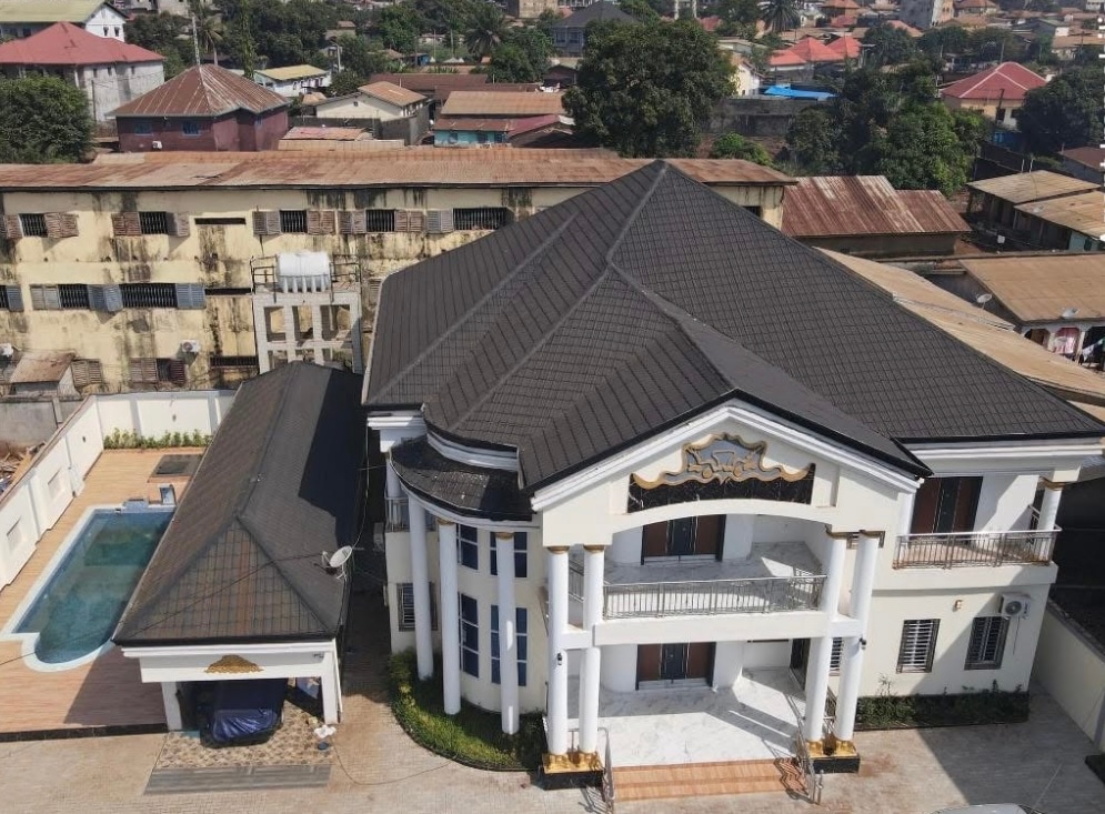 An aerial view showcases a spacious duplex highlighted by a striking facade with columns and a prominent gabled roof. The private pool is visible beside the house, enclosed by a tiled area, while surrounding green landscapes and urban rooftops create a contrast in the background.