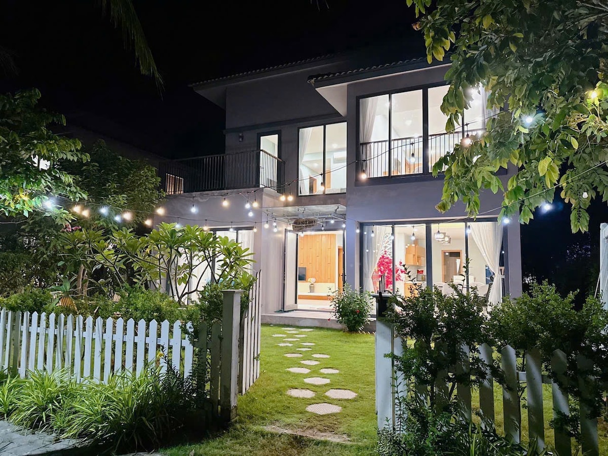 The exterior of a modern two-story house is illuminated at night, featuring large glass windows and a welcoming ambiance. Lush greenery and string lights create a festive atmosphere. A neatly arranged stone path leads to the entrance, bordered by a white picket fence.