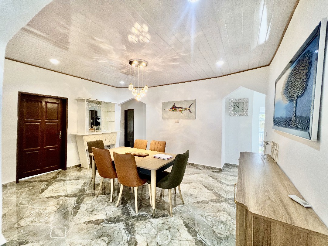 The dining area features a large table surrounded by six chairs, offering ample space for meals. A sideboard runs along one wall, with a decorative piece above it. Light fixtures illuminate the room's elegant marble flooring and white walls.