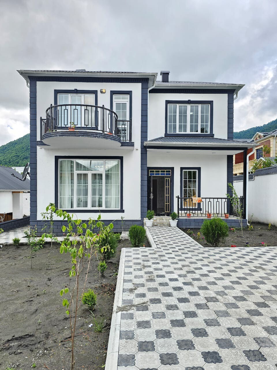 A two-story house features a modern façade, with white walls contrasted by dark trim. The entrance is framed by symmetrical balconies on each floor. A patterned pathway leads to the door, bordered by landscaped greenery and a pebbled area that enhances the outdoor space.