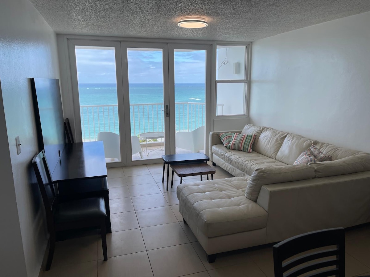 A bright living area is highlighted by large glass doors that open to a balcony with a view of the ocean. A light-colored sectional sofa offers ample seating, complemented by a dark wooden coffee table and a dining table nearby. The walls are painted a neutral white, enhancing the airy feel.