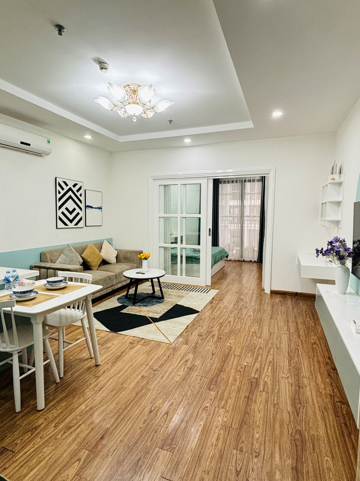 A living area is depicted, featuring a comfortable sofa and an inviting dining table set for meal preparation. Natural light enters through glass doors leading to an adjacent room, while decorative elements are arranged thoughtfully across the space, enhancing a sense of warmth and relaxation.
