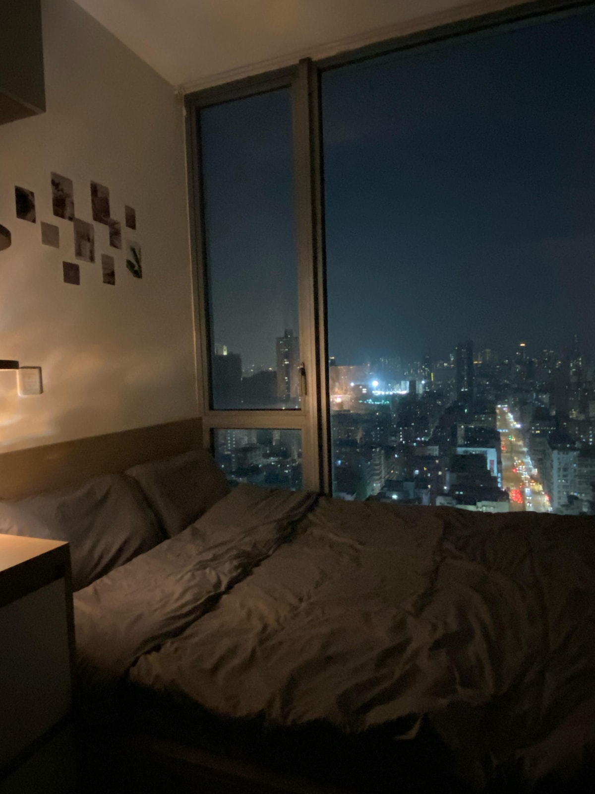 A spacious bed layered with soft grey bedding is positioned near a large window. A soft glow from a bedside lamp illuminates the room while a city skyline is visible through the glass, showcasing distant lights and a vibrant street below.