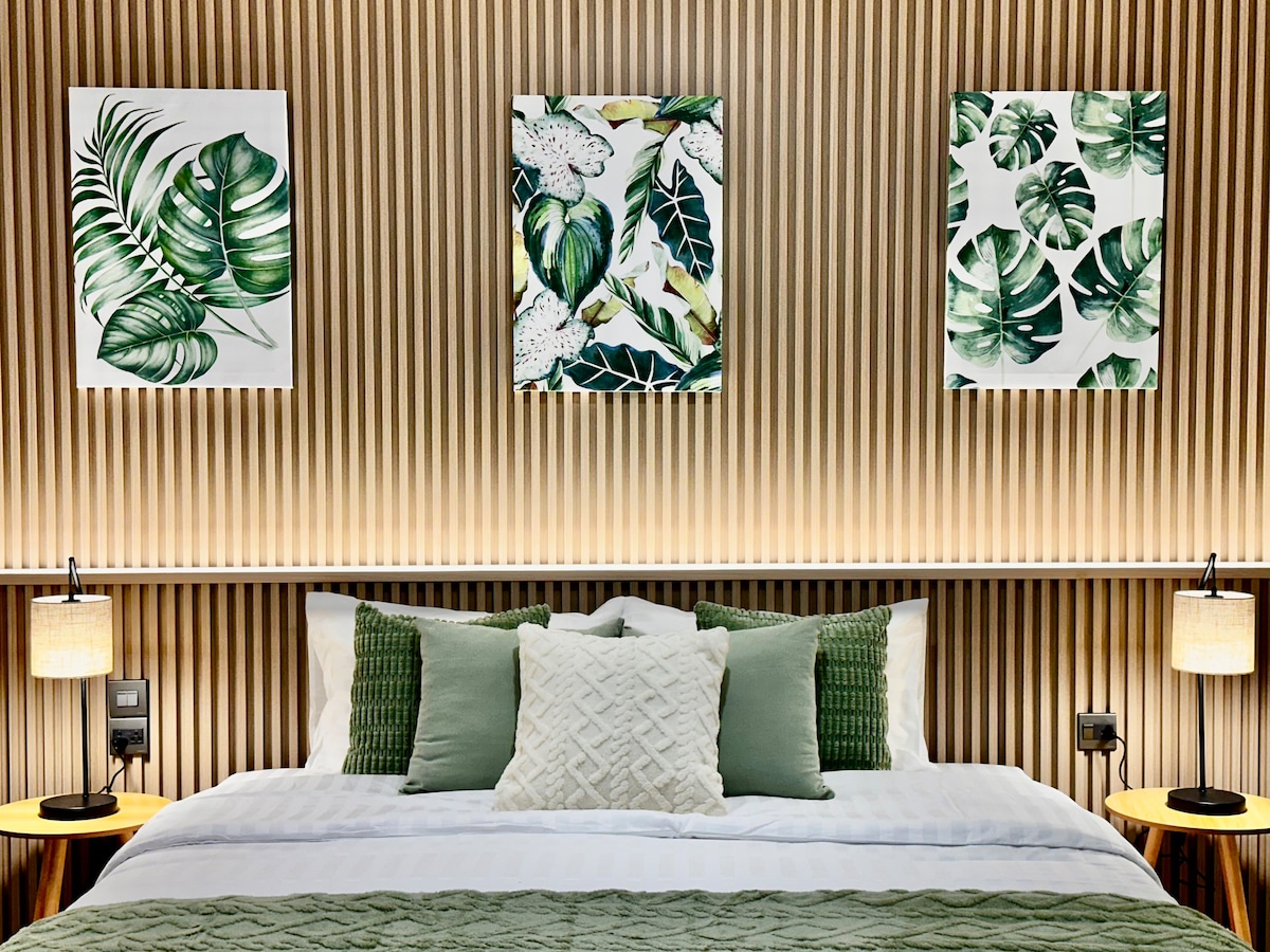 A cozy sleeping area features a king bed adorned with soft green accents and decorative pillows. Two bedside lamps provide gentle illumination. The wall showcases three nature-themed art pieces, while wood paneling complements the inviting atmosphere.