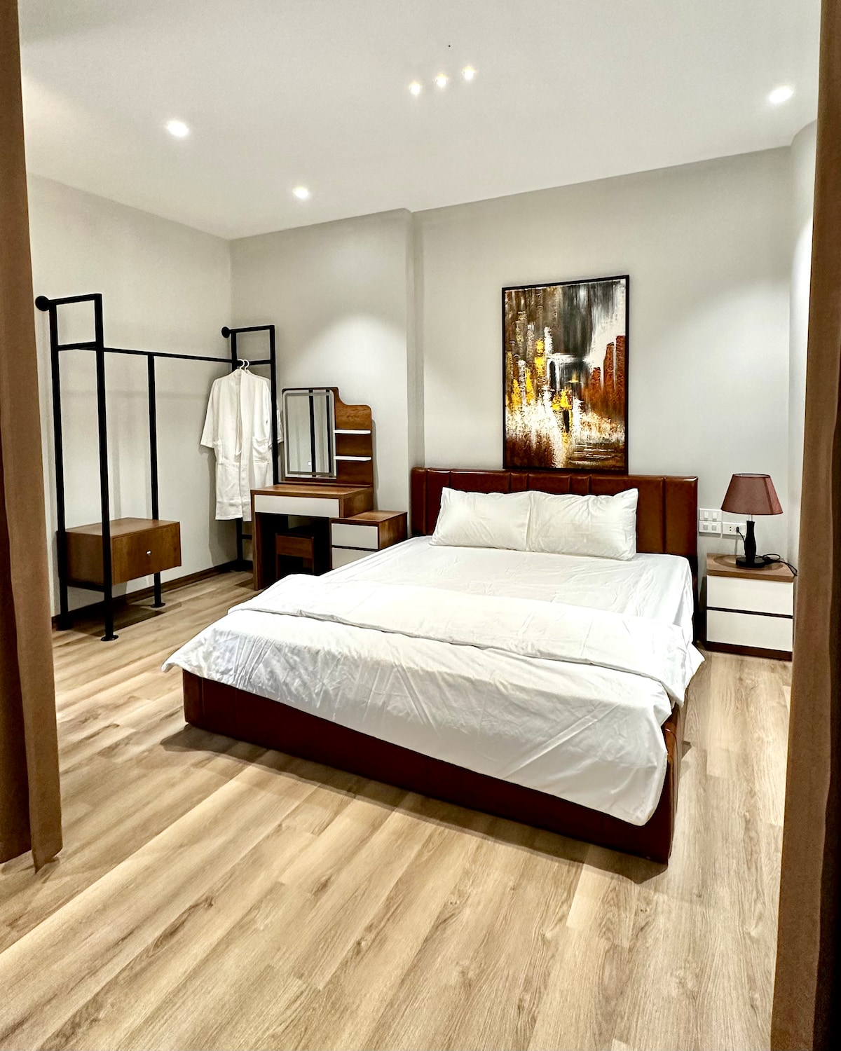 A cozy bedroom is equipped with a spacious bed dressed in white linens. A large piece of artwork adorns the wall above the bed. A functional closet and a mirror stand nearby, complemented by two bedside tables. Soft lighting creates a calm environment.