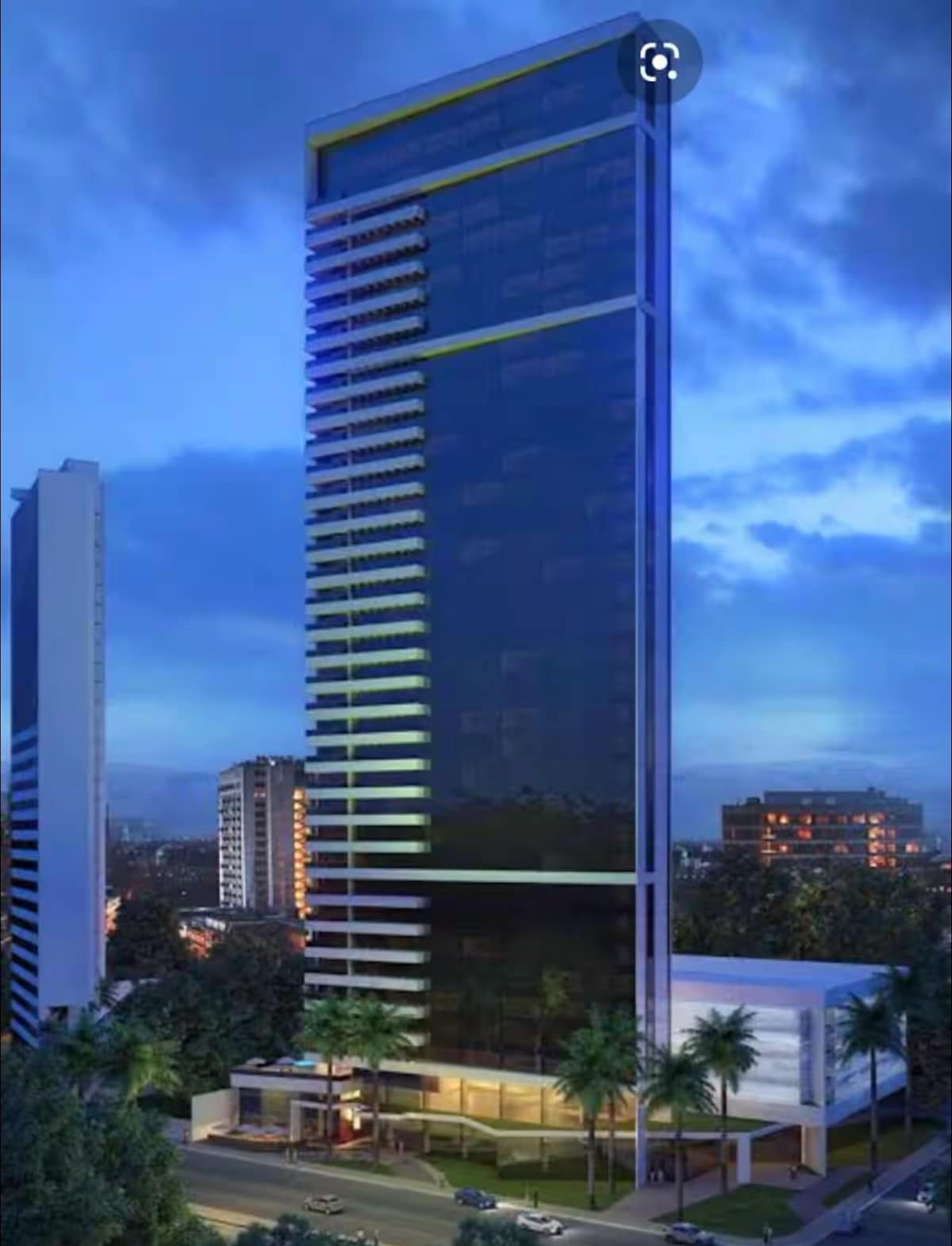 The modern high-rise building showcases a sleek glass façade reflecting the evening sky. Lush palm trees are positioned in the foreground, highlighting the landscaped entrance. The surrounding urban environment includes other structures, contributing to the contemporary cityscape.