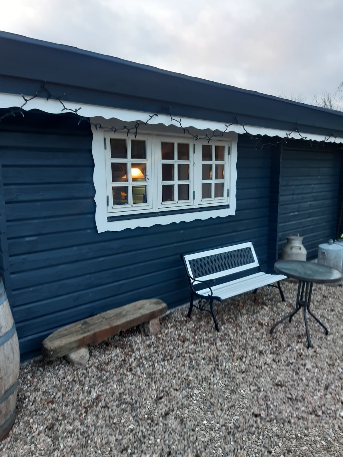 A charming exterior features a dark blue wooden structure with a white-framed window, highlighting the rustic design. An assortment of gravel surrounds the building, complemented by a wooden bench and a small stone table for outdoor seating.