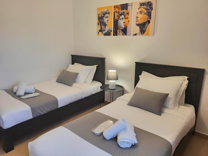 Udobna spavaća soba s dva kreveta: 2 udobna kreveta s tamnim uzglavljem, svježa bijela posteljina, sivi detalji, ručnici i papuče. Upečatljiva umjetnost nadahnuta klasikom, pogled na grad, tihi klima-uređaj, veliki ormar. Čisto, spokojno, moderno