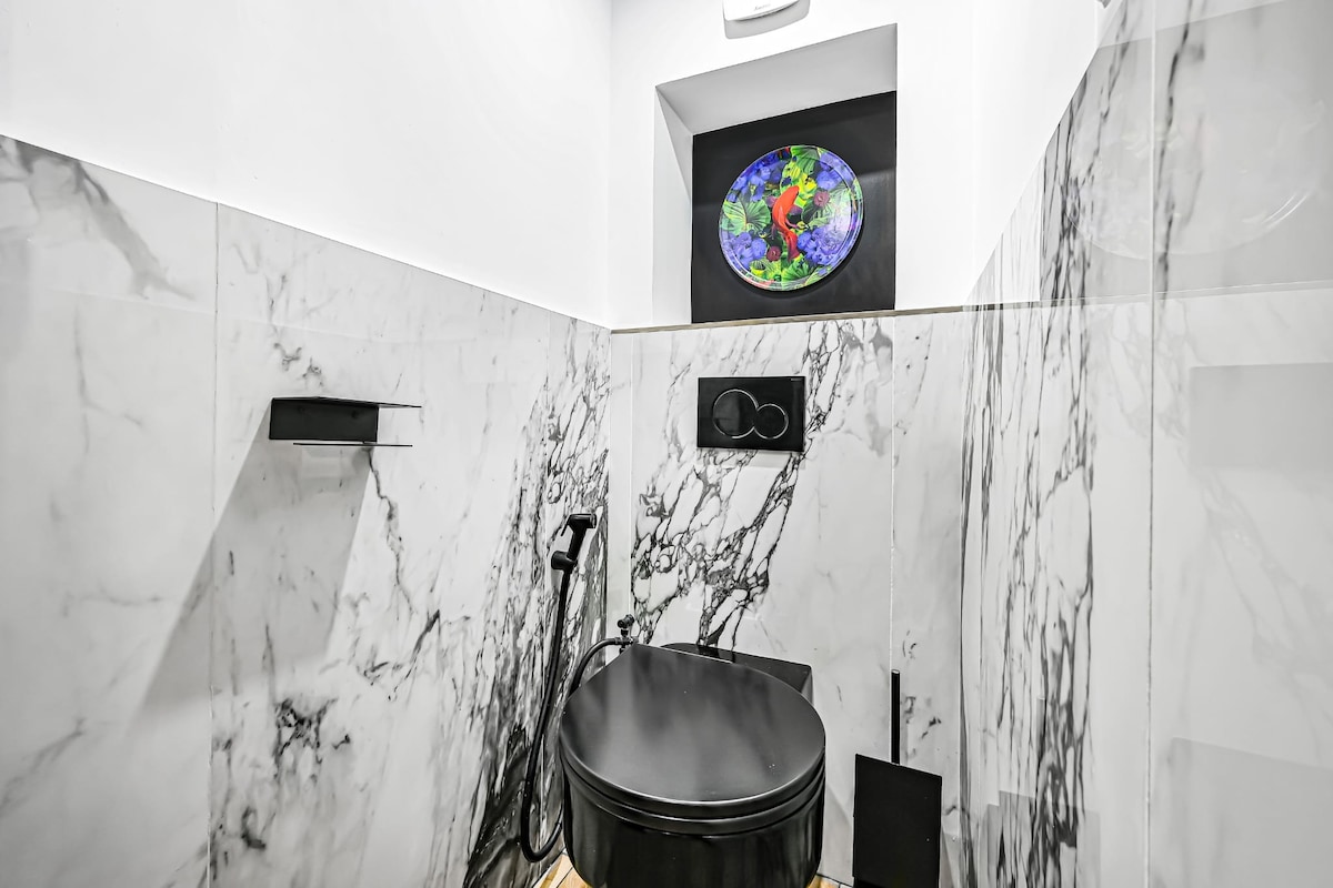 A modern bathroom is shown with marble walls featuring striking black and white veining. A sleek black toilet is positioned against the wall, and a colorful stained glass window adds a unique touch of character to the serene space.