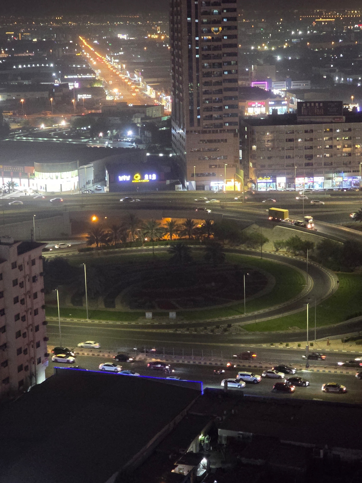 An aerial view captures a busy roundabout surrounded by palm trees. Vehicles are seen navigating the traffic, while illuminated buildings and streetlights create a vibrant cityscape in the background, highlighting the urban atmosphere during the evening.