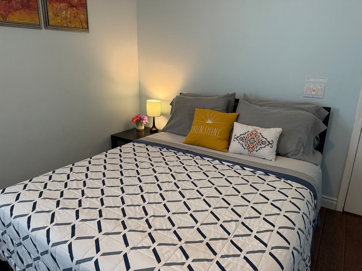 A cozy bedroom is presented with a spacious bed dressed in a patterned blanket. A small bedside lamp illuminates the space, accompanied by decorative pillows, including a bright yellow one with the word 'Sunshine.' Soft blue walls enhance the room's calming ambiance.
