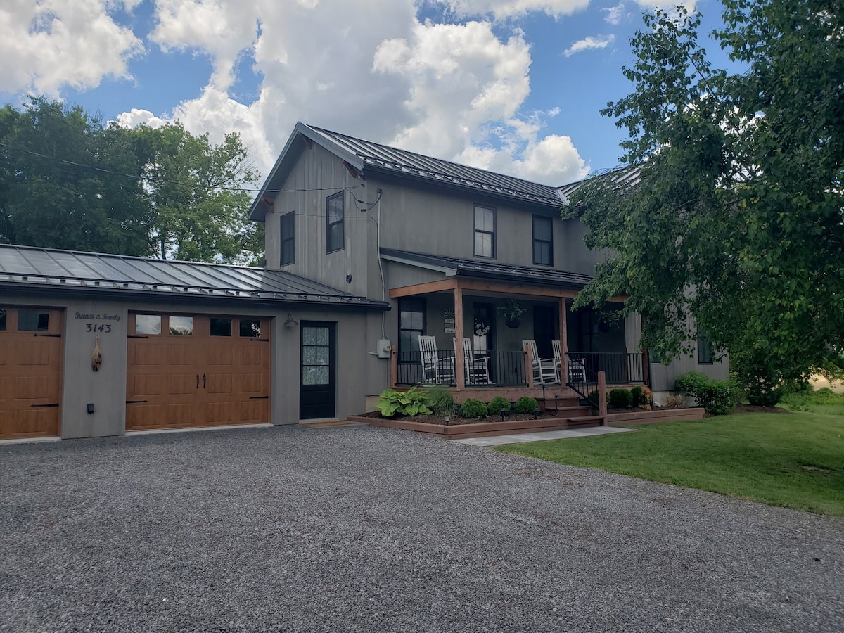 A modern farmhouse exterior is presented, featuring a combination of wooden and metal elements. A covered porch with rocking chairs invites relaxation. The gravel driveway leads up to the house, surrounded by lush greenery and a clear blue sky with scattered clouds.