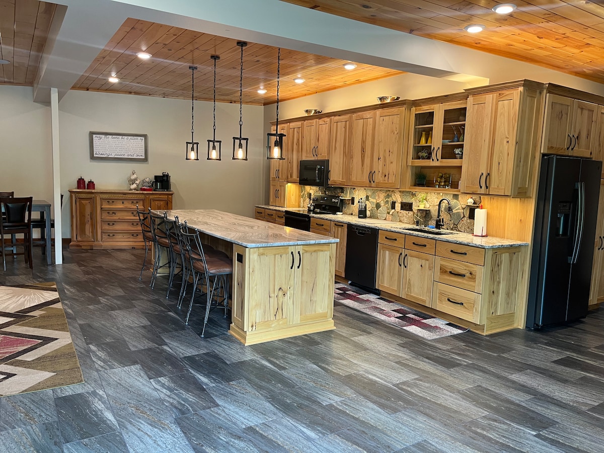 A spacious kitchen is highlighted by warm wooden cabinetry and stone countertops. The area features a large island with seating and modern appliances, including a refrigerator and oven. Ample lighting enhances the inviting workspace, with a dining table visible in the background.