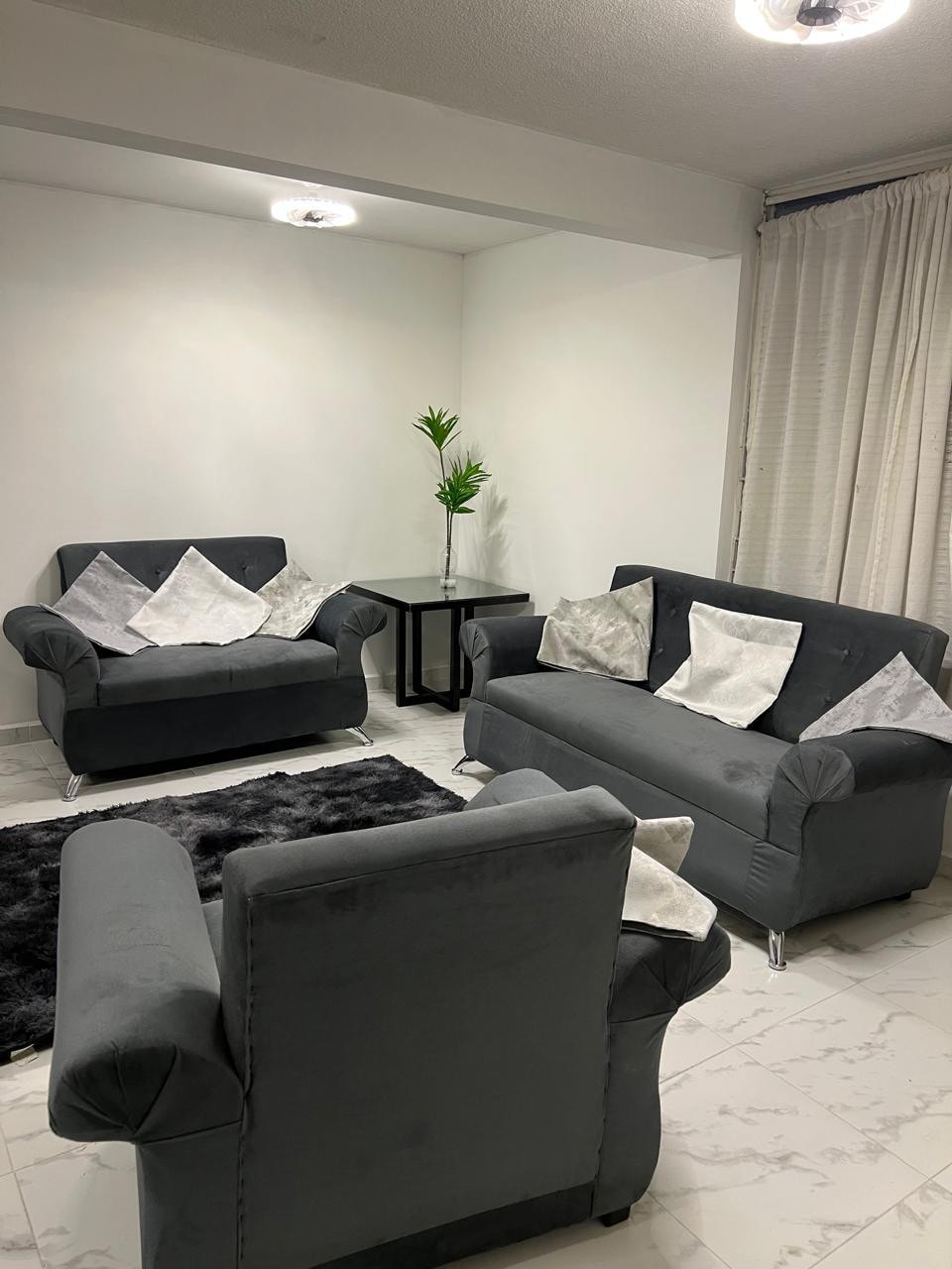 A modern living area is furnished with two grey couches and a pair of armchairs, all accented with decorative pillows. A small black table is positioned between the couches. Light-colored curtains frame the windows, and a plush area rug covers the floor.
