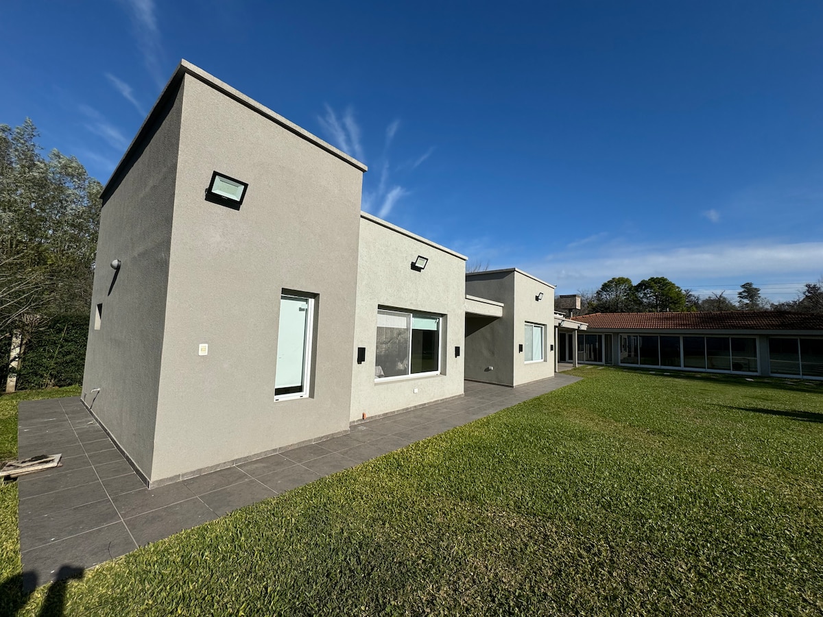 A modern two-story building is depicted with a sleek exterior in light gray. Large windows provide ample natural light, and a spacious patio area is visible, surrounded by well-kept green grass. A clear blue sky enhances the overall openness of the space.