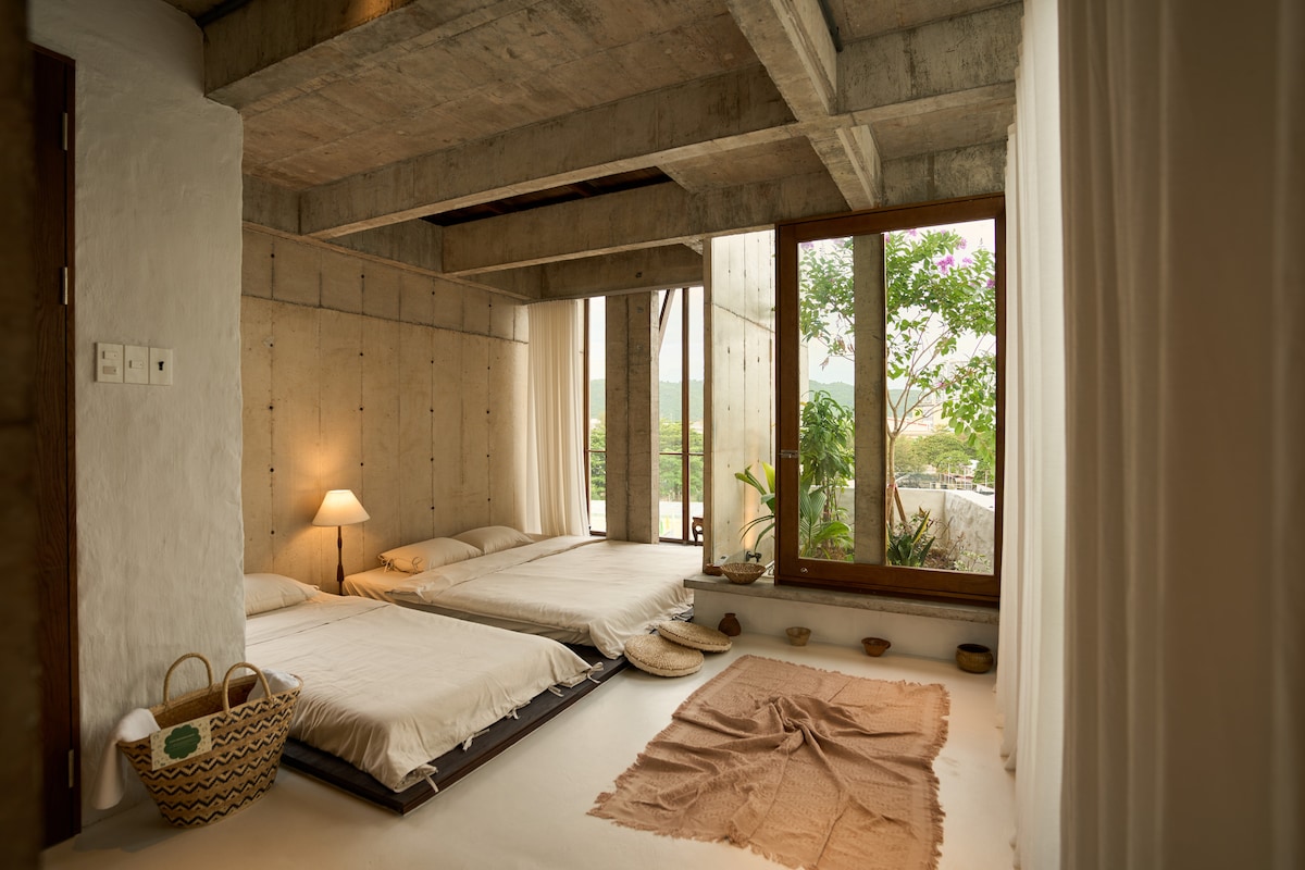 A serene bedroom features minimalist decor, with two spacious beds positioned thoughtfully against a textured concrete wall. Natural light filters through large windows, illuminating the room's simplicity. A lamp offers gentle illumination, and potted plants enhance the connection to nature.