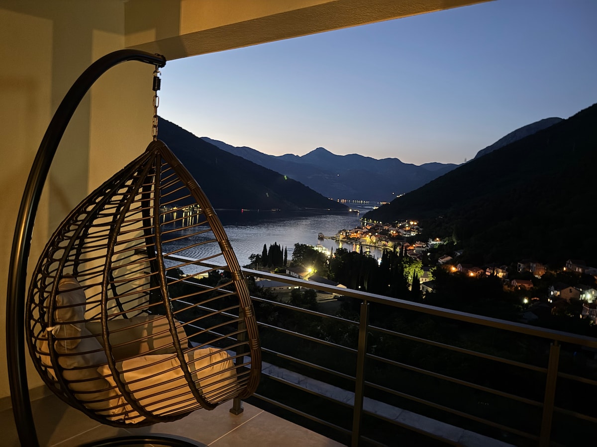 A hanging chair is positioned on a balcony, offering a serene view of the illuminated bay at dusk. Mountains rise in the background, while the warm lights of a nearby town reflect on the water, creating a peaceful atmosphere.