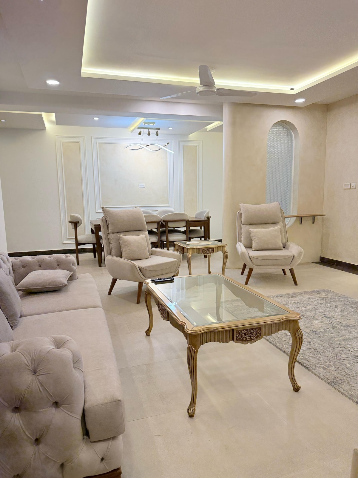 A modern living area features a tufted beige sofa complemented by two matching armchairs. A glass coffee table is positioned in the center, while a subtle area rug adds texture. A dining table is visible in the background, surrounded by solid wooden chairs.