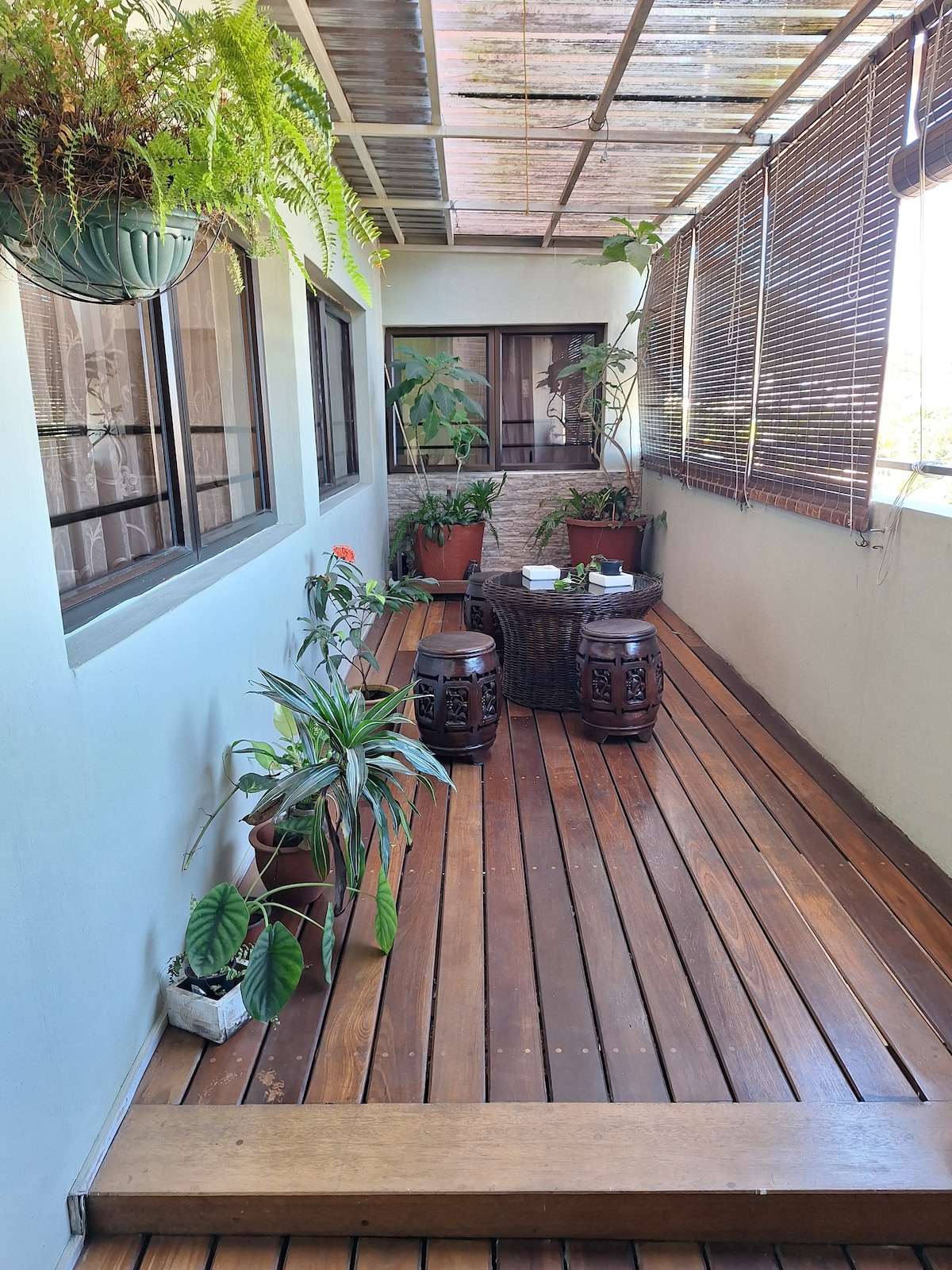 A covered balcony features wooden flooring and several potted plants. Two decorative stools are placed beside a small round table. Natural light filters through the window blinds, creating a serene atmosphere perfect for relaxation.