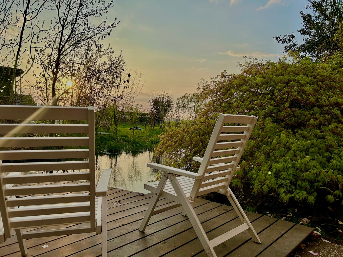 A wooden deck features two white lounge chairs positioned to overlook a serene pond. The setting sun casts a warm glow over the landscape, surrounded by greenery and soft, blooming plants. Gentle reflections of the trees and sky can be seen in the water.