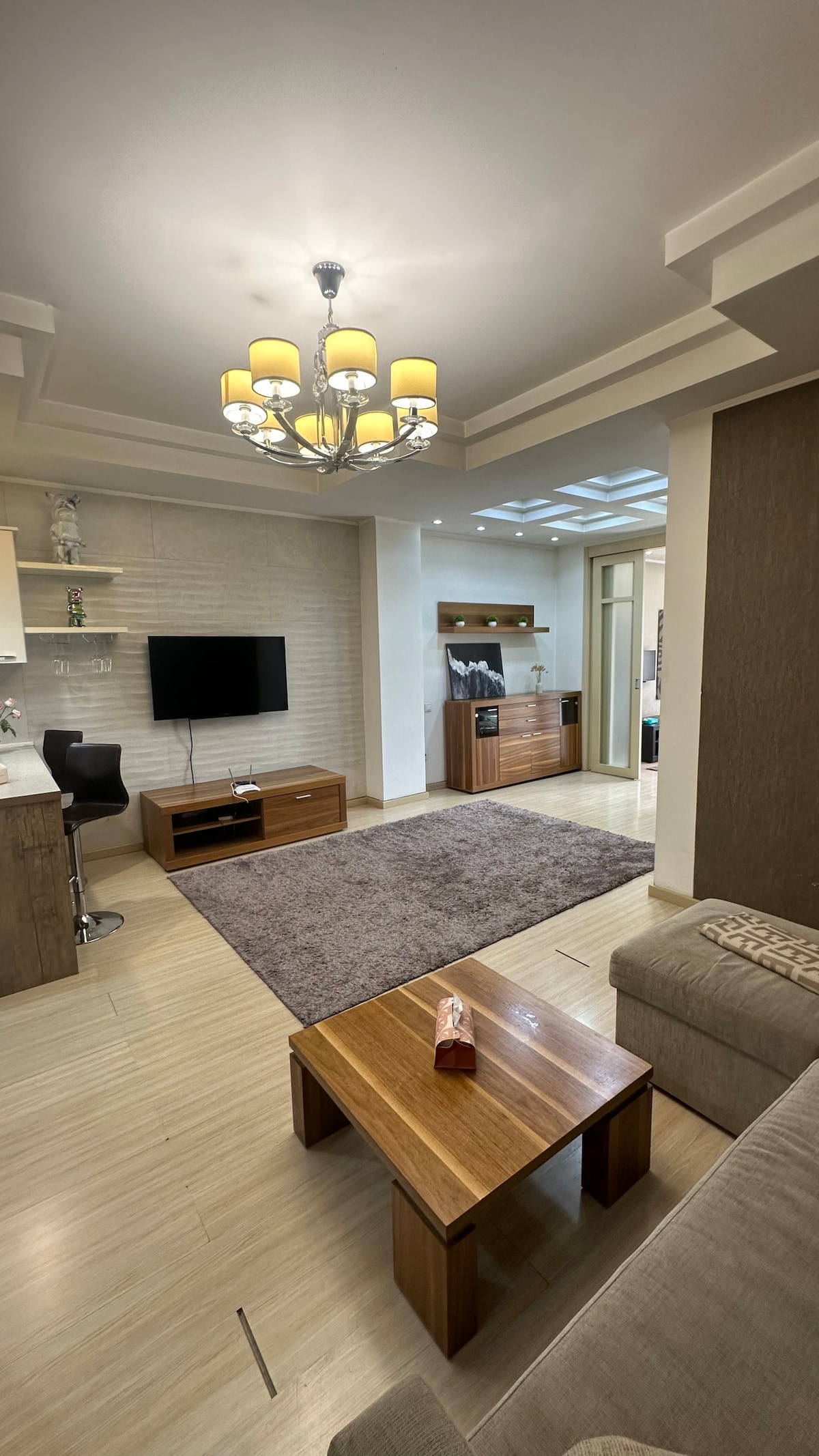 A stylish living area features a soft rug and a wooden coffee table. A comfortable sofa is positioned nearby, while a television is mounted on the wall. Natural light is enhanced by a modern chandelier, creating a welcoming space near the kitchen area.