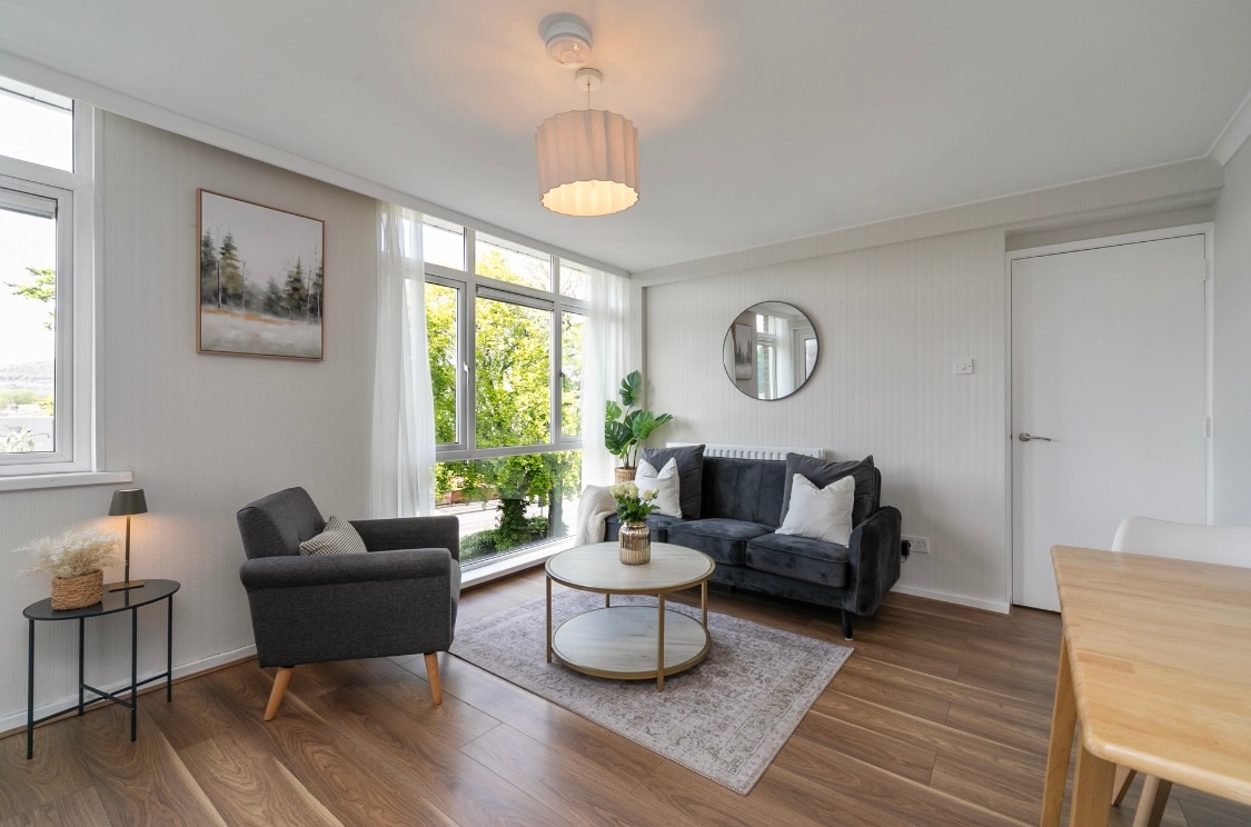 A cozy living area includes a dark sofa, a grey armchair, and a round coffee table positioned on a soft rug. Sunlight filters through large windows, illuminating the space. A plant and artwork add a touch of nature, while a door leads to another room.